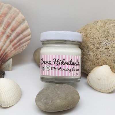 Frasco de creme hidratante em vidro com rótulo rosa e branco, colocado entre pedras e conchas