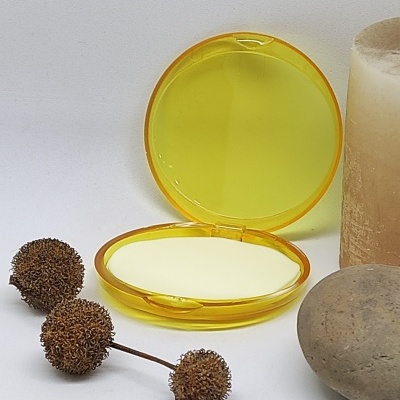 Estojo redondo amarelo translúcido aberto com espelho e base branca ao lado de pedras e flores secas