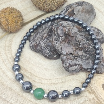 Pulseira de contas metálicas prateadas com conta verde central sobre madeira, casca e elementos naturais secos