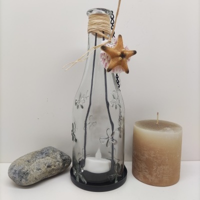 Garrafa de vidro decorada com flor em relevo e corda, estrela-do-mar, concha, pedra cinzenta e vela bege sobre superfície branca