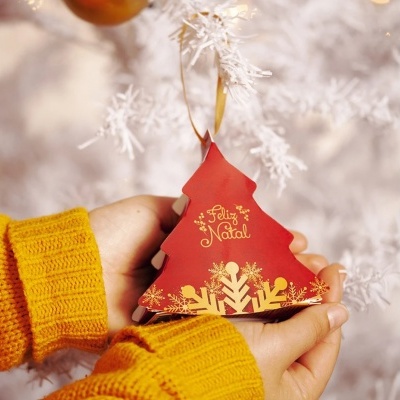 Ornamento de Natal em forma de árvore vermelha com texto 'Feliz Natal' segurado por duas mãos com camisolas amarelas