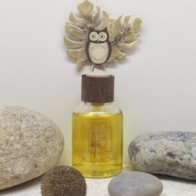 Frasco de difusor de aroma com tampa decorada por coruja, rodeado por pedras sobre superfície clara