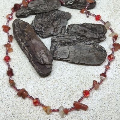 Colar de pedras naturais vermelhas, castanhas e contas vermelhas em superfície bege com casca de árvore.