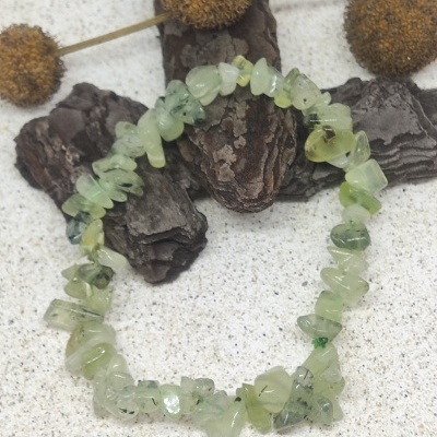 Pulseira de pedras verdes translúcidas com textura irregular em fundo cinza com casca de árvore e flores secas