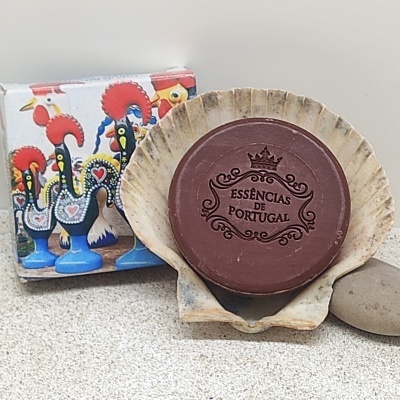 Sabonete 'ESSÊNCIAS DE PORTUGAL' numa concha ao lado de embalagem com galos de Barcelos coloridos e pedra