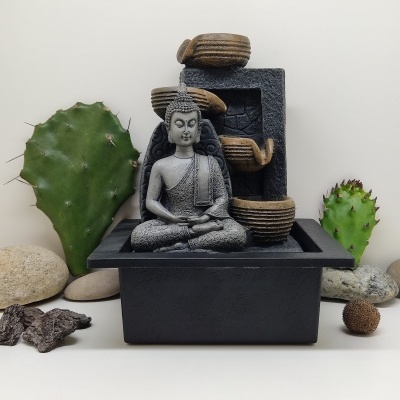 Fonte decorativa com estátua do Buda e tigelas castanhas, base preta, rodeada por pedras e cactos