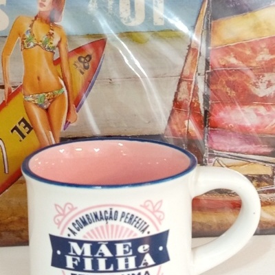 Caneca branca com interior rosa e texto em português sobre mãe e filha, em frente a imagem de mulher com prancha de surf