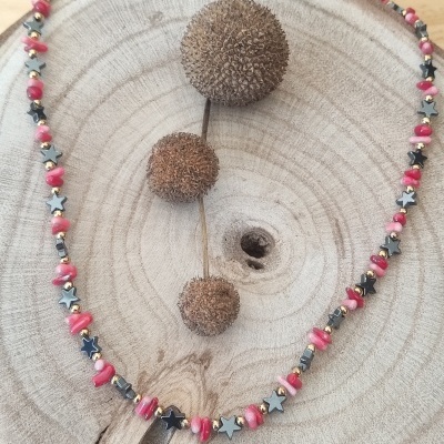 Colar com contas rosa, estrelas cinzentas e bolinhas douradas sobre superfície de madeira com sementes secas