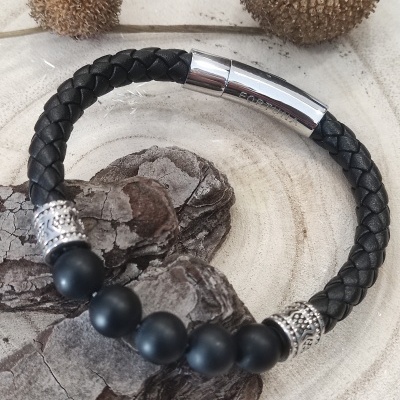 Pulseira preta em couro trançado com contas negras e detalhes prateados sobre casca e fundo claro