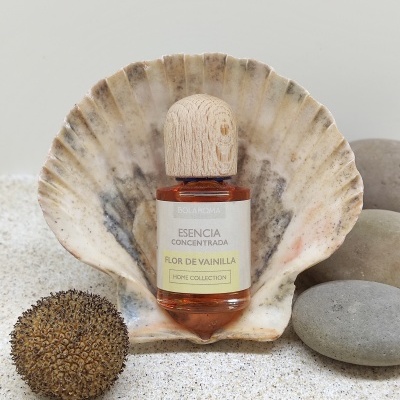 Frasco de essência Flor de Vainilla num cenário natural com concha, pedras e areia