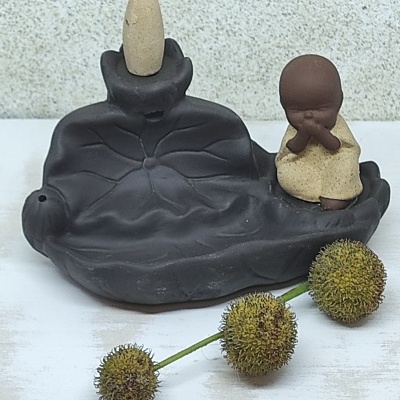 Incensário cerâmico em forma de folha com monge e cone de incenso, decorado com flores esféricas.