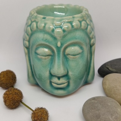 Vela aromática em cerâmica azul esverdeada com formato de cabeça de Buda rodeada por pedras e flores secas