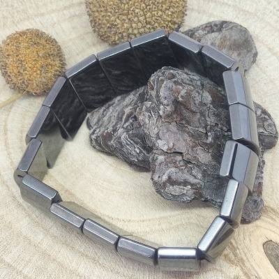 Pulseira metálica cinza escuro com peças retangulares sobre madeira e sementes secas