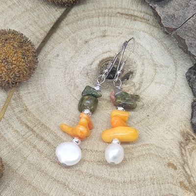 Brincos com contas verdes, laranja e pérolas sobre madeira com elementos naturais secos.