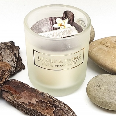 Vela aromática em copo de vidro fosco com etiqueta dourada ao centro, rodeada por casca de madeira e pedras lisas