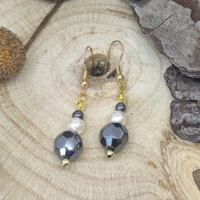 Brincos com contas facetadas azul metálico, pérolas brancas e esferas douradas em fundo de madeira