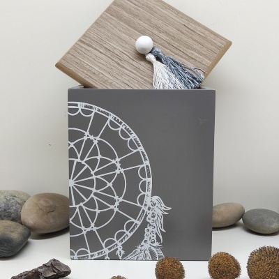 Caixa decorativa cinzenta com design de mandala branco e tampa de madeira clara com pingente, rodeada por pedras e galhos secos.
