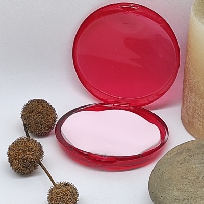 Compacto vermelho aberto com pó branco, bolbos secos, vela e pedra sobre fundo branco.