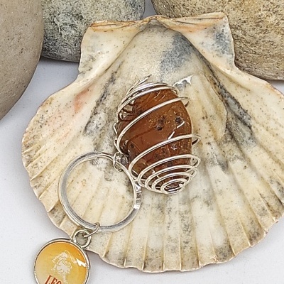 Chaveiro com pedra castanha em fio de metal e pendente amarelo com texto LEO, numa concha, pedras ao fundo