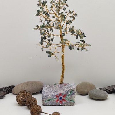 Árvore decorativa de fios dourados e pedras verdes numa base de pedra pintada com flor, rodeada por pedras naturais e ramos secos.