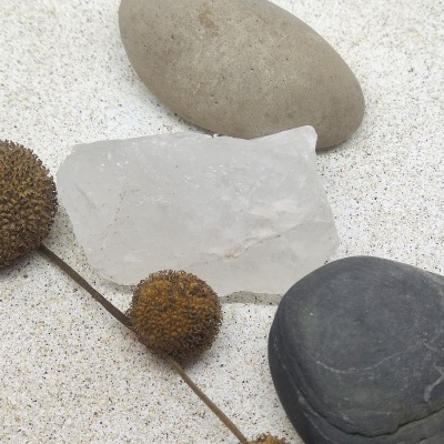 Três pedras lisas em tons bege, branco e preto com um ramo seco com bolas castanhas em fundo branco granuloso