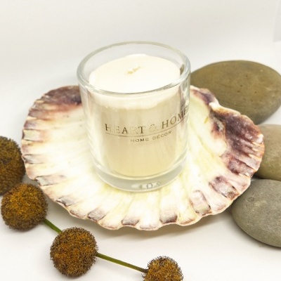 Vela branca em copo de vidro com texto dourado sobre concha natural, pedras lisas e bolas secas.