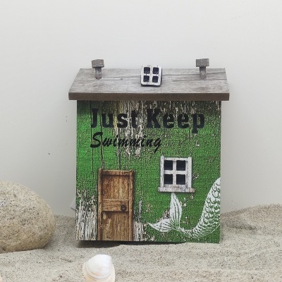 Caixa decorativa em forma de casa verde com texto 'Just Keep Swimming' sobre areia com conchas