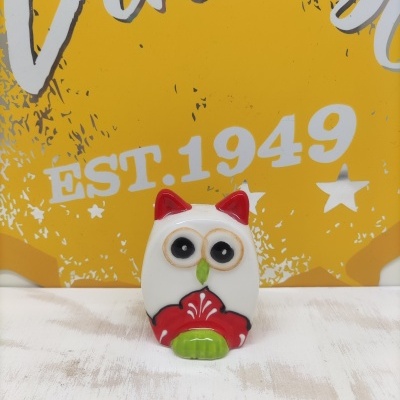 Figura decorativa de coruja em cerâmica com detalhes vermelhos e verdes diante de fundo amarelo com texto EST. 1949