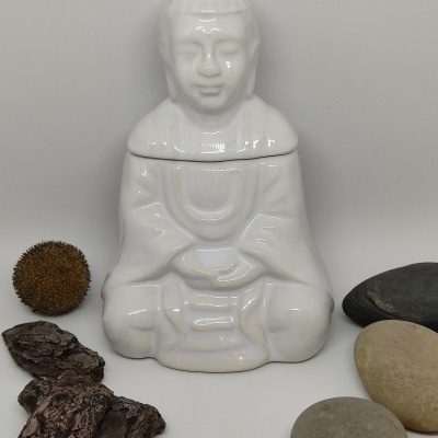 Escultura branca de figura meditativa com pedras e elemento vegetal seco ao redor.