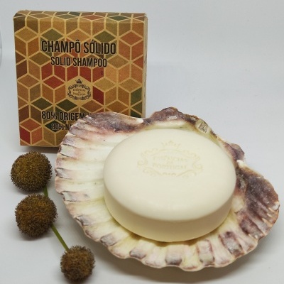 Champô sólido creme numa concha com embalagem geométrica atrás e flores castanhas ao lado