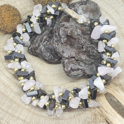 Colar de pedras rosa claro, pretas e esferas douradas sobre tronco de madeira com sementes e pedaço de madeira