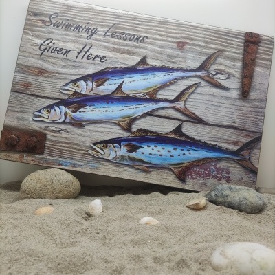 Placa decorativa de madeira com peixes pintados e texto 'Swimming Lessons Given Here' sobre areia com pedras