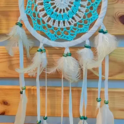 Apanhador de sonhos em crochet azul e branco com penas e contas verdes pendurado numa parede de madeira.