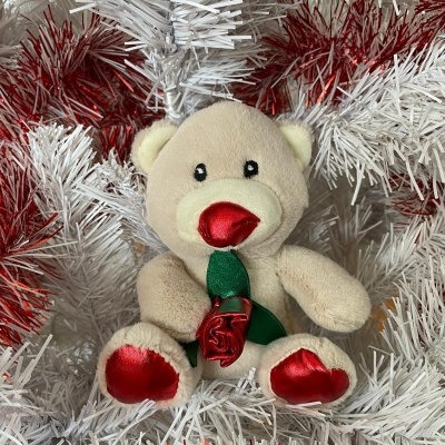 Ursinho de peluche bege com rosa vermelha segurada sentado em guirlanda branca e vermelha.