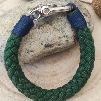 Pulseira trançada verde com fecho metálico prateado e detalhes azuis sobre pedra e madeira