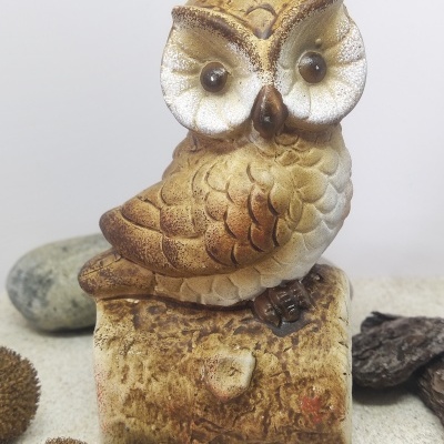 Figura decorativa de coruja em tons castanhos e beges sobre bloco que imita madeira.