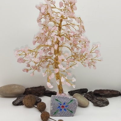 Árvore decorativa de fios dourados e pedras rosa com base quadrada decorada, em fundo branco com pedras e madeira
