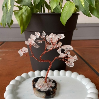 Árvore decorativa com cristais em base de pedra sobre superfície branca, vaso preto e planta ao fundo.