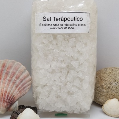 Pacote transparente de sal terapêutico grosso branco com etiqueta, rodeado por conchas e pedra sobre fundo branco.