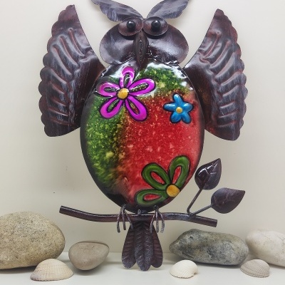 Escultura decorativa de coruja em metal colorido e castanho com flores sobre fundo com pedras e conchas.