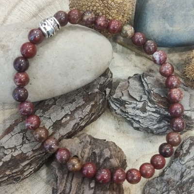 Pulseira de contas vermelhas, castanhas e bege com detalhe metálico, sobre casca de árvore e pedras