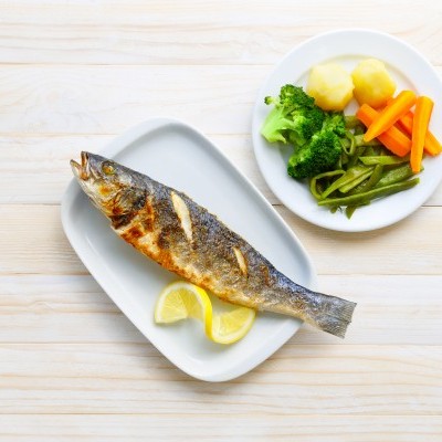Peixe grelhado com fatias de limão e prato com vegetais cozidos sobre mesa de madeira clara