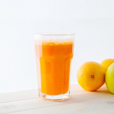 Copo de sumo laranja com frutas e legumes ao lado numa mesa de madeira clara