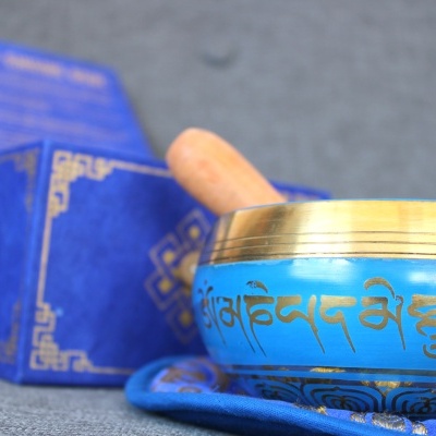 Tigela de som azul com inscrições douradas e bastão de madeira sobre almofada azul, com caixa azul decorativa ao fundo.