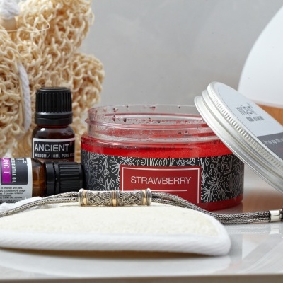 Produtos para cuidados pessoais incluindo esfoliante de morango, óleos essenciais, esponja de banho e difusor de aroma sobre tampo branco.