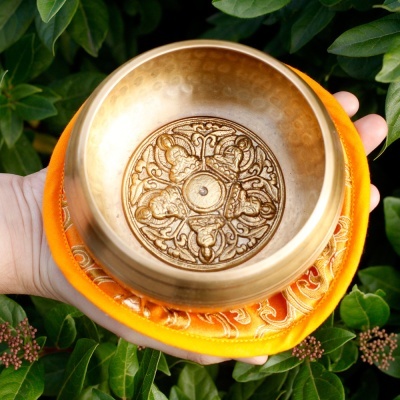 Tigela tibetana dourada em metal com almofada laranja e detalhes dourados sobre fundo de folhas verdes