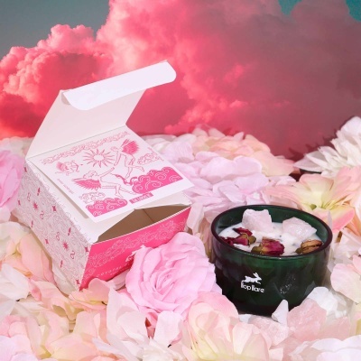 Caixa branca aberta e vela preta com pétalas em fundo de nuvens cor-de-rosa.