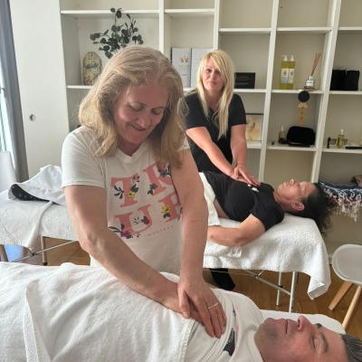 Sala com duas macas de massagem, duas mulheres a fazer massagens a duas pessoas, estante branca ao fundo com produtos e plantas