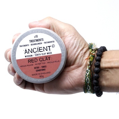 Pote metálico com máscara de barro vermelho ANCIENT, segurado por mão com pulseiras, fundo branco
