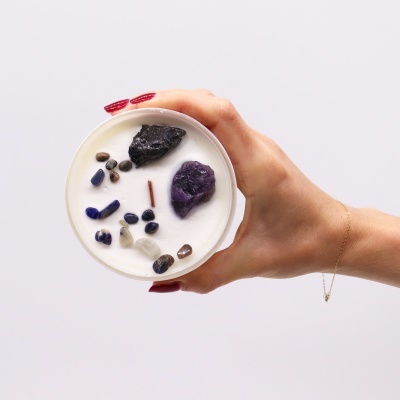 Vela branca com pedras naturais coloridas na superfície sendo segurada por uma mão com unhas vermelhas